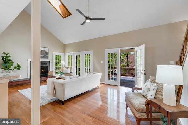 living room with a skylight, light wood-style floors, high vaulted ceiling, a ceiling fan, and a lit fireplace