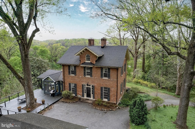 colonial-style house featuring a wooden deck, roof with shingles, a chimney, brick siding, and a wooded view