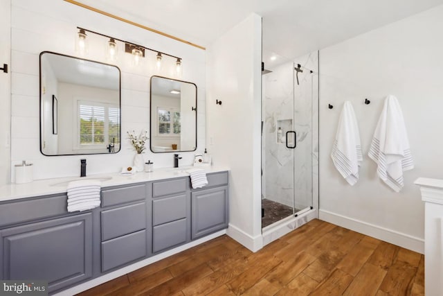 full bathroom with wood finished floors, double vanity, a sink, baseboards, and a marble finish shower