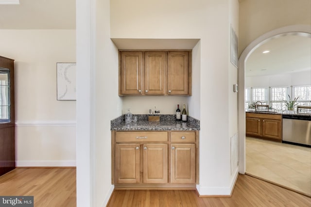 bar featuring stainless steel dishwasher, light wood-style flooring, a sink, arched walkways, and baseboards