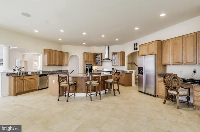 kitchen featuring brown cabinetry, an island with sink, a breakfast bar area, wall chimney exhaust hood, and appliances with stainless steel finishes