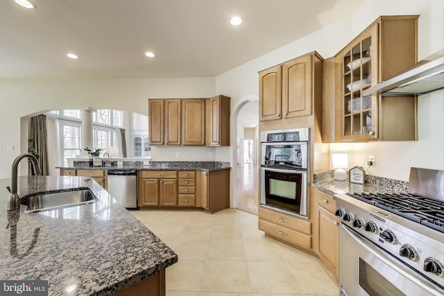 kitchen with wall chimney range hood, dark stone countertops, a sink, arched walkways, and stainless steel appliances