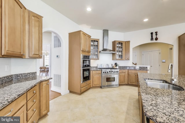 kitchen with stainless steel appliances, wall chimney range hood, a sink, arched walkways, and stone counters