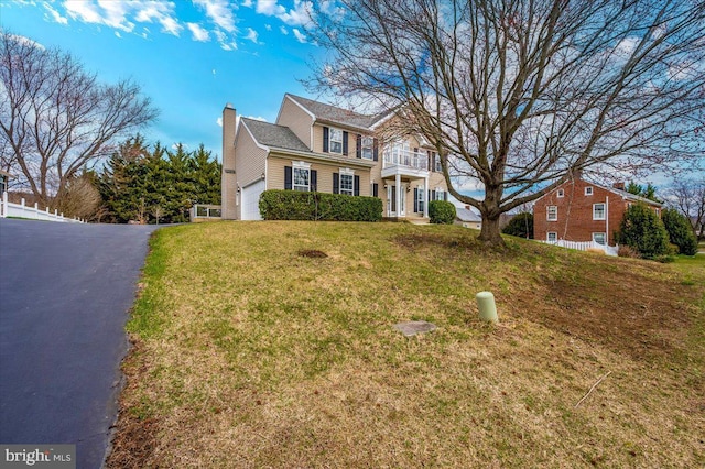 colonial house featuring a balcony, a chimney, driveway, an attached garage, and a front yard