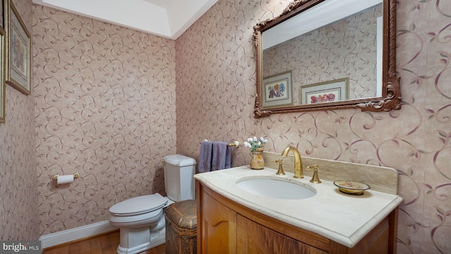 half bath with wallpapered walls, wood finished floors, toilet, and vanity