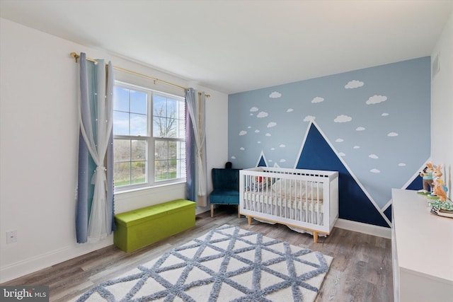 bedroom featuring a nursery area, an accent wall, wood finished floors, wallpapered walls, and baseboards