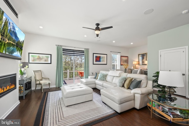 living area featuring baseboards, ceiling fan, a glass covered fireplace, and wood finished floors