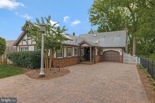 tudor-style house featuring decorative driveway, fence, a garage, stucco siding, and brick siding