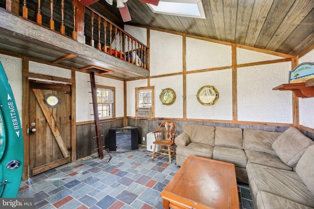 living room with vaulted ceiling with skylight, wainscoting, wooden walls, cooling unit, and ceiling fan