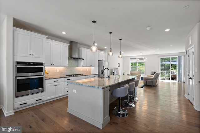 kitchen featuring white cabinets, open floor plan, wall chimney exhaust hood, a kitchen island with sink, and appliances with stainless steel finishes