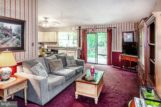 living room featuring wallpapered walls, a textured ceiling, carpet, and a ceiling fan
