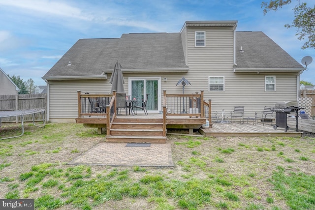 rear view of property with fence, roof with shingles, a trampoline, and a deck