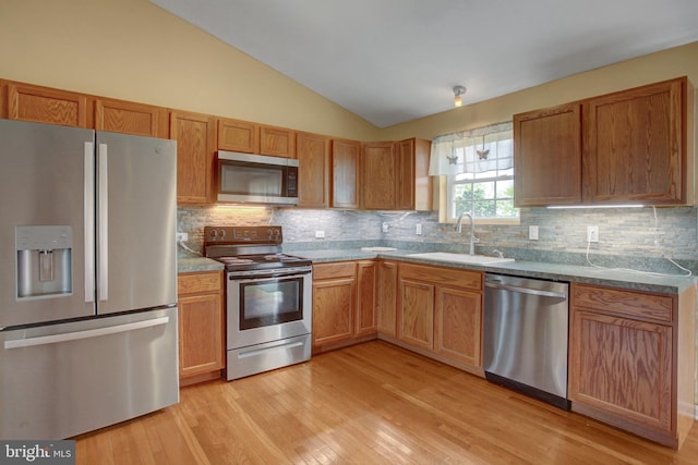 kitchen featuring appliances with stainless steel finishes, a sink, vaulted ceiling, light wood finished floors, and backsplash