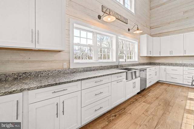 kitchen featuring stainless steel dishwasher, a sink, white cabinets, wood walls, and light stone countertops