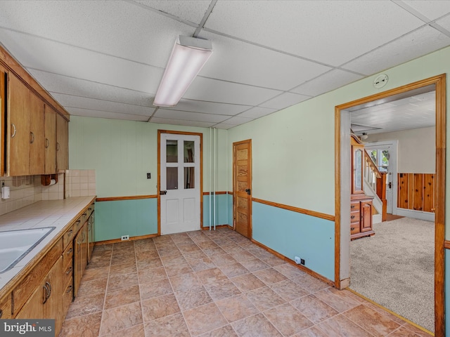 kitchen with brown cabinetry, baseboards, wooden walls, tile counters, and a paneled ceiling