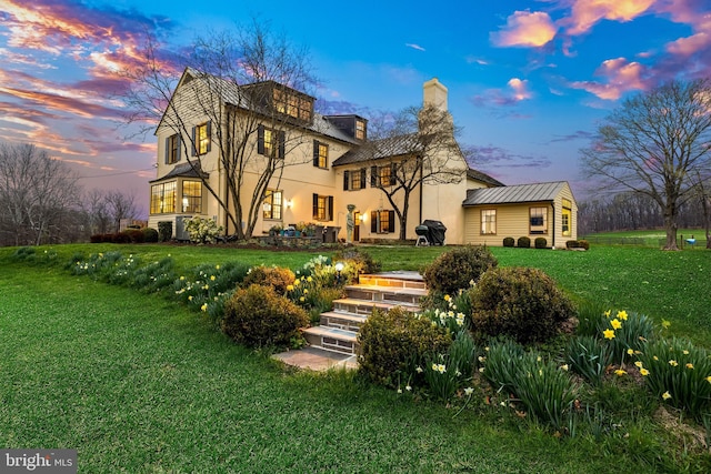 back of house at dusk featuring a standing seam roof, a yard, stucco siding, and a chimney