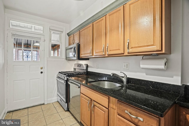 kitchen with brown cabinets, stainless steel appliances, a sink, light tile patterned flooring, and dark stone counters