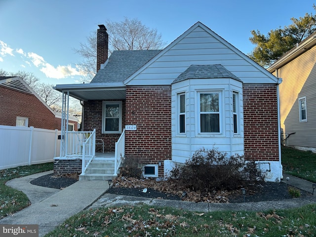 bungalow-style house featuring covered porch, a chimney, fence, brick siding, and roof with shingles