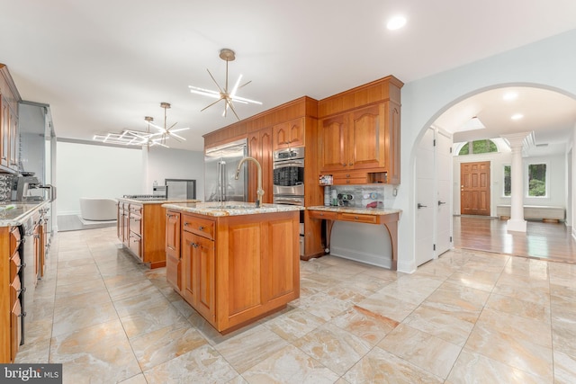 kitchen with decorative columns, a center island with sink, arched walkways, stainless steel appliances, and brown cabinetry