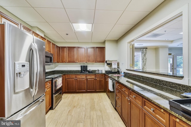 kitchen with dark stone counters, brown cabinets, light wood-style floors, appliances with stainless steel finishes, and a paneled ceiling