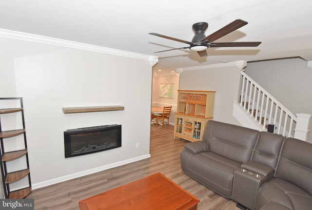 living area with ceiling fan, a glass covered fireplace, stairs, wood finished floors, and ornamental molding