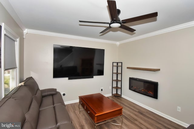 living area with ornamental molding, a glass covered fireplace, wood finished floors, baseboards, and a ceiling fan