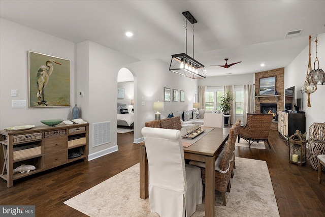 dining area featuring arched walkways, a stone fireplace, dark wood-type flooring, a ceiling fan, and recessed lighting