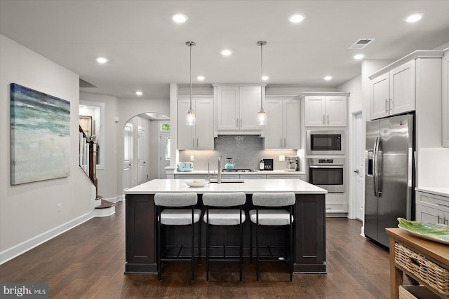 kitchen featuring appliances with stainless steel finishes, arched walkways, light countertops, dark wood-type flooring, and an island with sink
