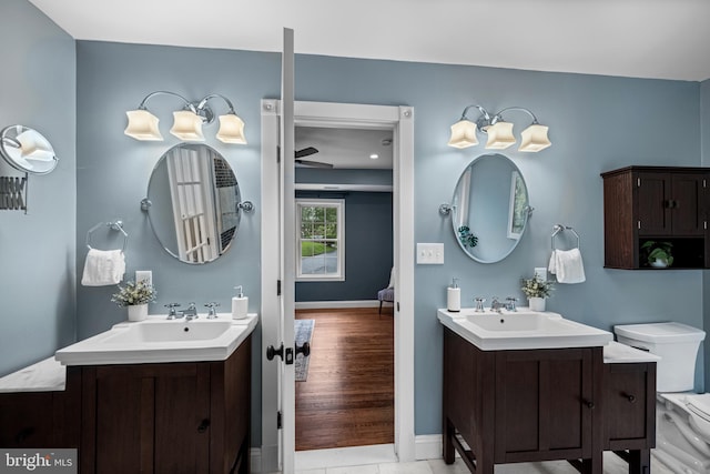 full bathroom featuring two vanities, baseboards, toilet, and ceiling fan