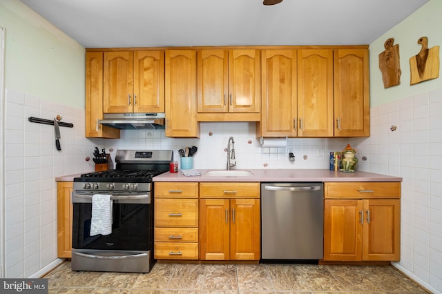 kitchen with tile walls, under cabinet range hood, brown cabinets, a sink, and appliances with stainless steel finishes