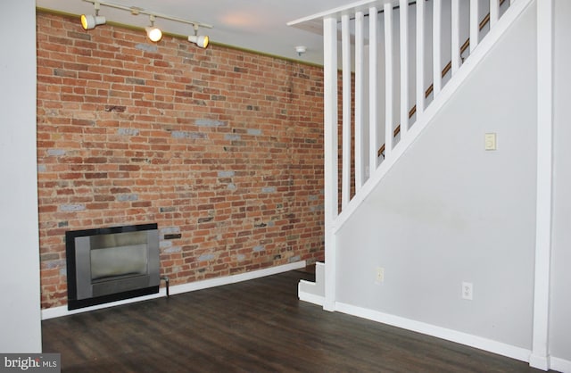 unfurnished living room featuring track lighting, baseboards, stairs, wood finished floors, and brick wall