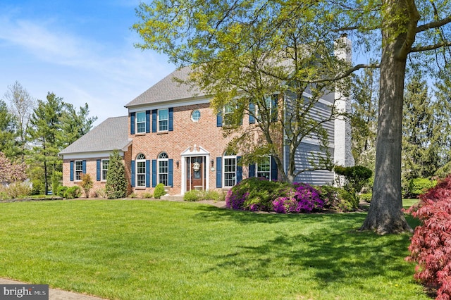 colonial inspired home with brick siding, a shingled roof, a front lawn, and a chimney