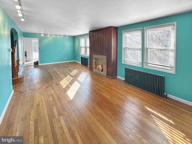 unfurnished living room featuring radiator, arched walkways, wood-type flooring, rail lighting, and a fireplace