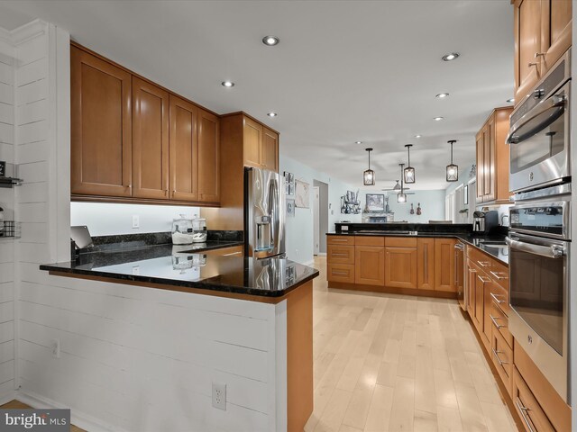 kitchen with appliances with stainless steel finishes, a peninsula, light wood-style floors, dark stone counters, and brown cabinets