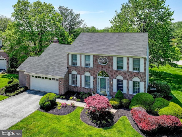 colonial home with roof with shingles, brick siding, a garage, a chimney, and driveway