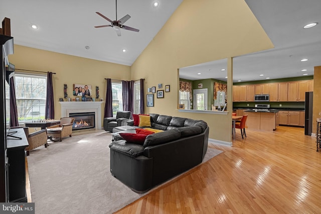 living room with a ceiling fan, recessed lighting, light wood-style flooring, high vaulted ceiling, and a glass covered fireplace