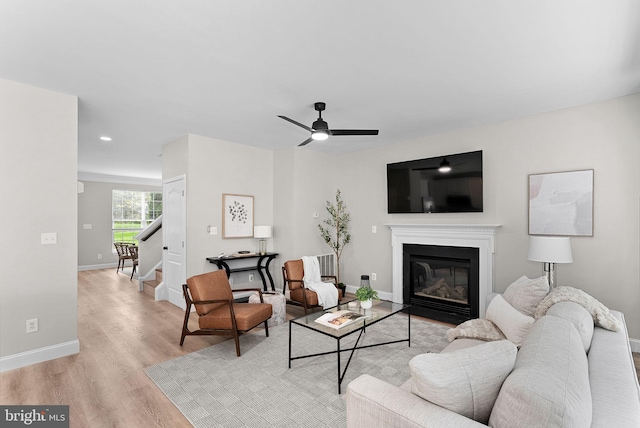 living area featuring baseboards, ceiling fan, stairs, light wood-style flooring, and a glass covered fireplace