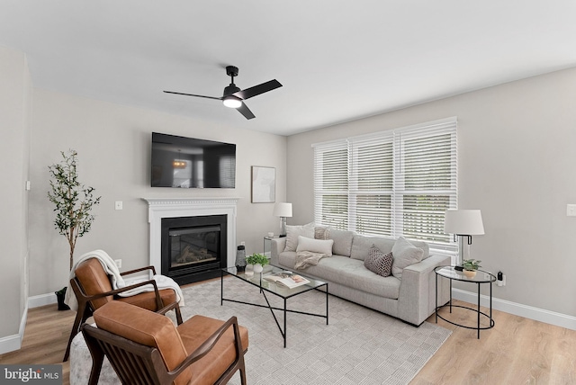 living area featuring ceiling fan, light wood-type flooring, a glass covered fireplace, and baseboards