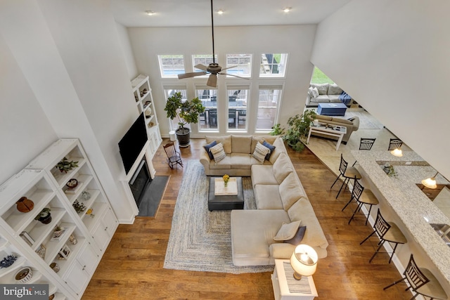 living room with ceiling fan, wood finished floors, a towering ceiling, and a fireplace with flush hearth