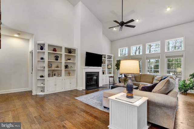 living area featuring baseboards, ceiling fan, wood finished floors, a fireplace with flush hearth, and high vaulted ceiling