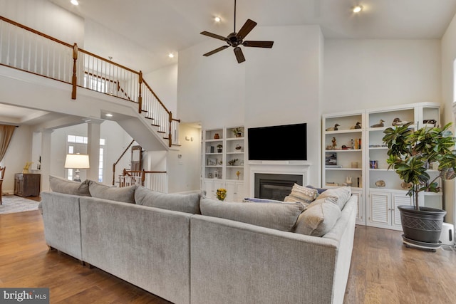living room with a towering ceiling, a ceiling fan, a glass covered fireplace, wood finished floors, and stairs