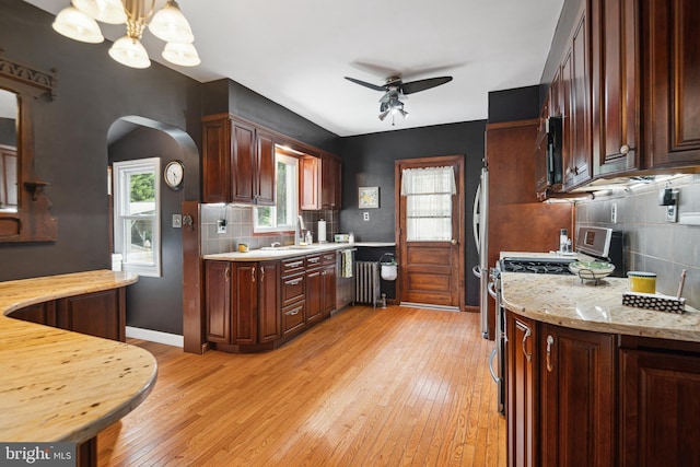 kitchen featuring backsplash, arched walkways, light wood-type flooring, stainless steel gas stove, and ceiling fan with notable chandelier