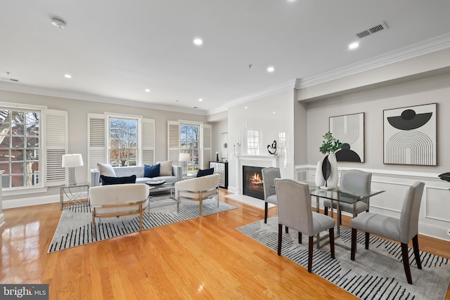 dining area featuring crown molding, a warm lit fireplace, light wood finished floors, visible vents, and recessed lighting