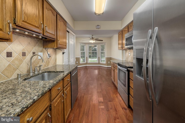 kitchen with brown cabinetry, dark wood-type flooring, dark stone counters, a sink, and stainless steel appliances