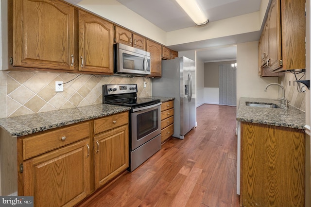 kitchen featuring appliances with stainless steel finishes, a sink, brown cabinetry, wood finished floors, and tasteful backsplash