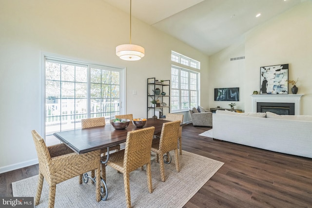 dining space with dark wood-style floors, high vaulted ceiling, a glass covered fireplace, visible vents, and baseboards