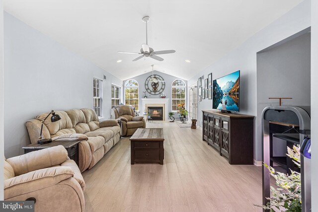 living room with a ceiling fan, a glass covered fireplace, vaulted ceiling, and light wood-style flooring