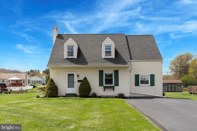 cape cod-style house with an outdoor structure, a front yard, stucco siding, roof with shingles, and a storage unit