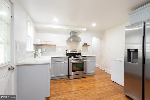 kitchen with stainless steel appliances, a sink, wall chimney range hood, gray cabinets, and open shelves