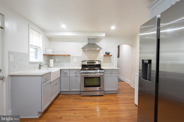 kitchen with light wood finished floors, open shelves, wall chimney range hood, gray cabinetry, and appliances with stainless steel finishes